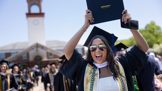 A graduate in reflective sunglasses in front of the Quinnipiac clocktower cheers with her diploma