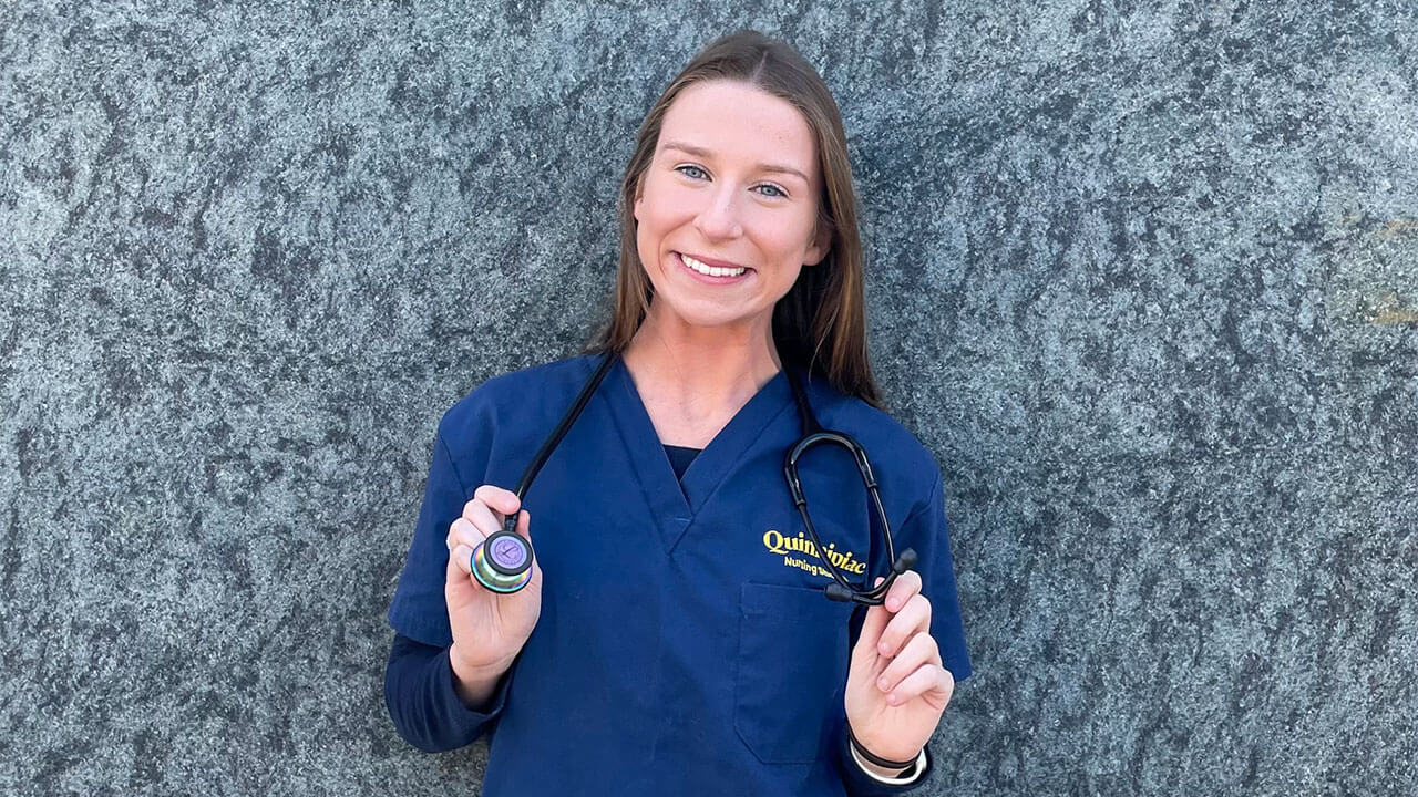 kaitlin gorman, a girl with light brown hair and blue eyes, smiles as she poses for the camera in her Quinnipiac nursing scrubs with a stethoscope around her neck