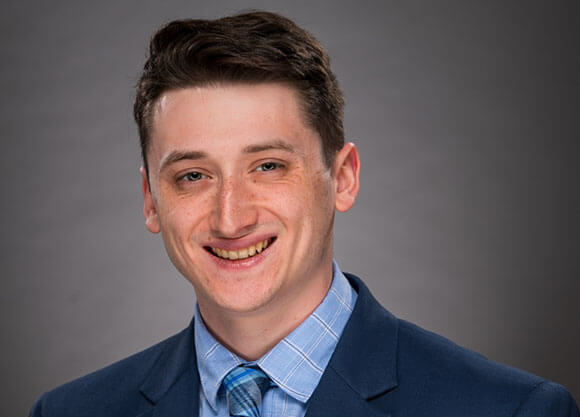 jack theall poses in a gray suit and light blue shirt, with a quinnipiac plaid tie in front of a light gray background. he has dark brown hair and blue eyes
