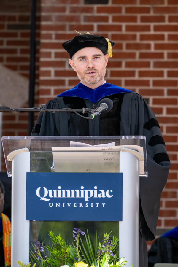 Eric Marrapodi wears Commencement robes and stands at a Quinnipiac podium on the library steps