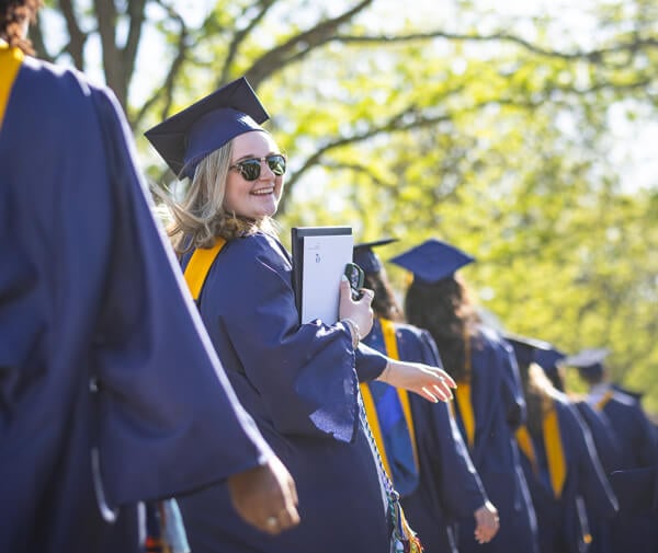 A graduate looks over her shoulder and smiles while walking in a line at Quinnipiac Commencement