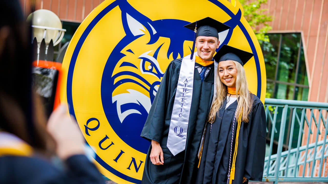 Two graduates smile in front a Quinnipiac Bobcat sign