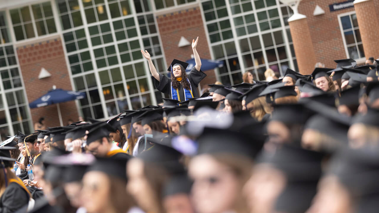 A graduate stands up above a sea of classmates to wave