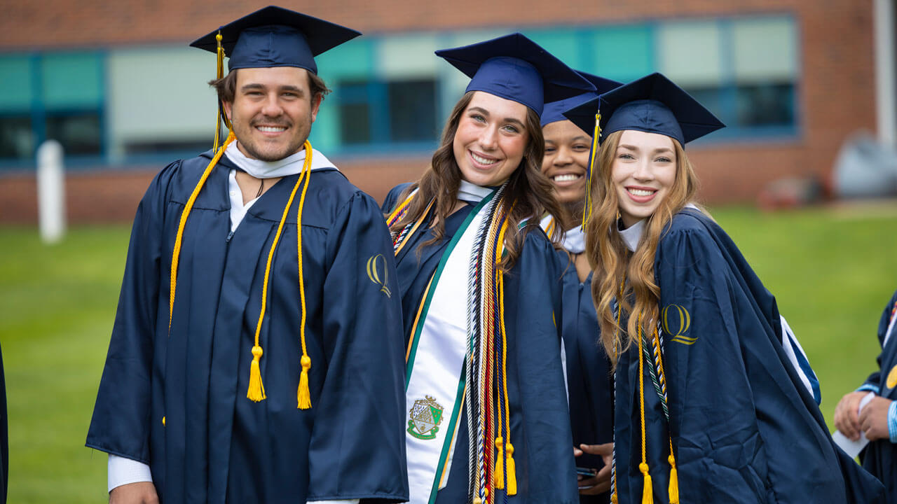 Four graduates stand close together and smile for a photo on the quad