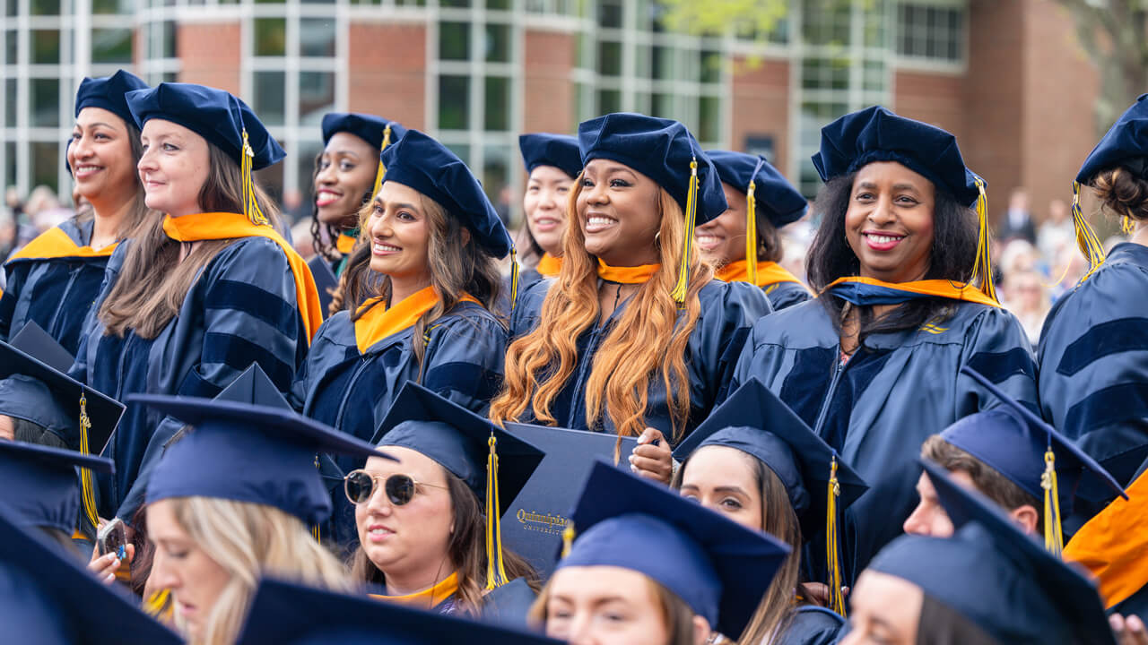 Half a dozen nursing graduates stand and smile in a row among a sea of graduates